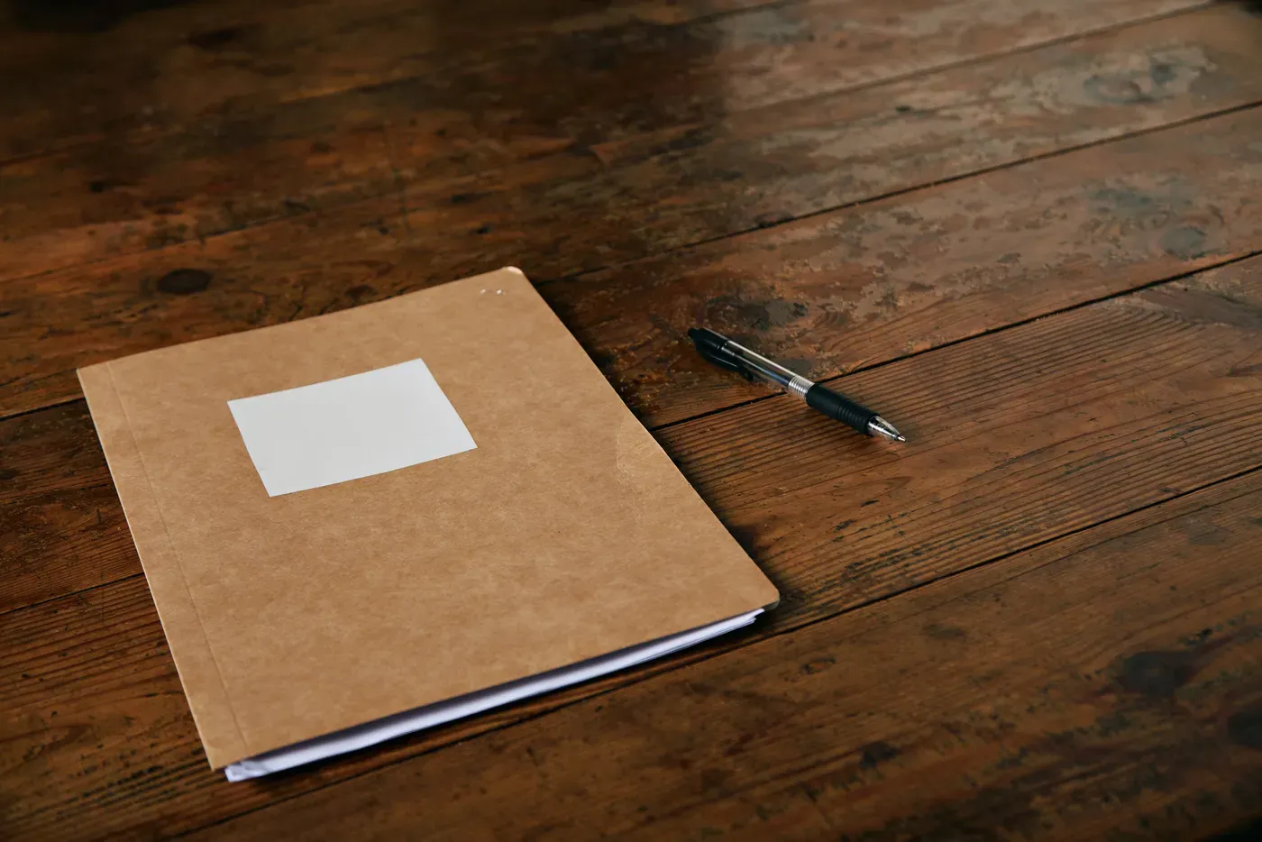 Pasta de papel pardo fechada, com uma etiqueta em branco, e uma caneta sobre uma mesa de madeira escura