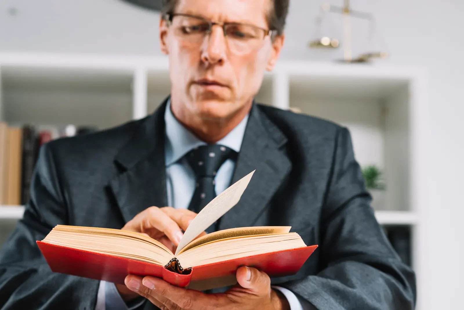 Homem de terno e óculos lendo um livro de capa vermelha, com uma balança da Justiça ao fundo